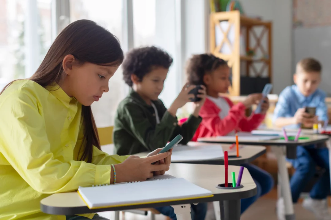 Crianças em uma sala de aula, concentradas enquanto exploram jogos educativos em um celular.